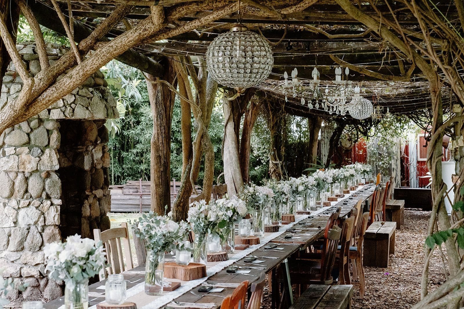 Long wedding banquet table styled with floral posies and rustic tables and chairs with chandeliers and fairy lights under long 20m arbour covered in vines