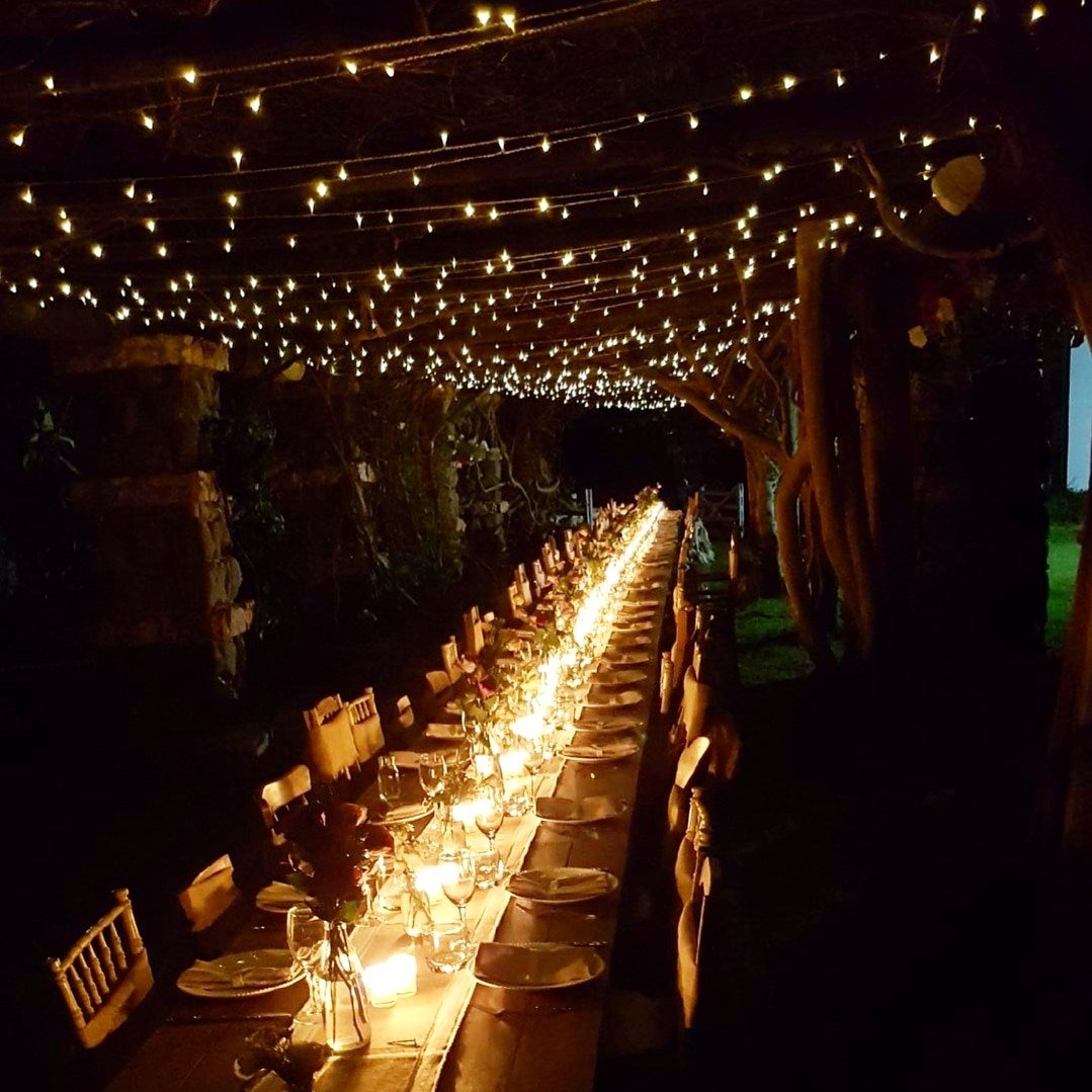 Long rustic wooden feasting table & chairs under canopy of vines & fairy lights hanging from the historic garden arbour covered in flowers Tables lit by candles