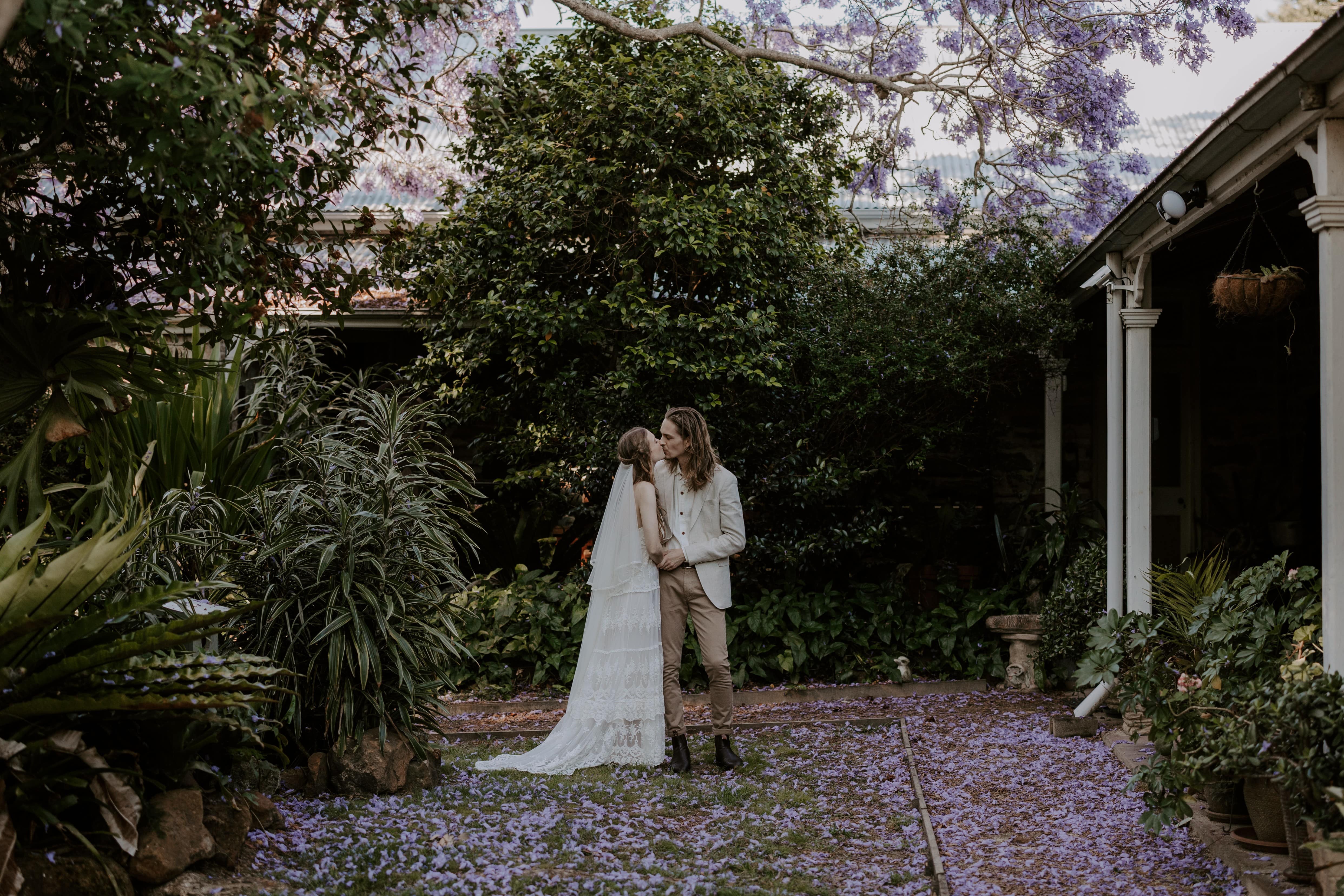 Bride and groom kissing in Tanilba House garden, Port Stephens, with jacarandah falling around them and on the ground like a carpet. Location Port Stephens
