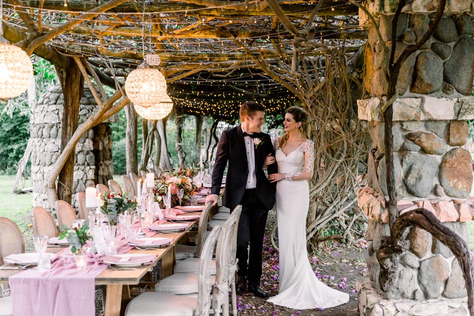 Bride and groom under rustic garden stone arbour with long banquet table set with elegant pink toned styling plus fairy lights and vintage chandeliers